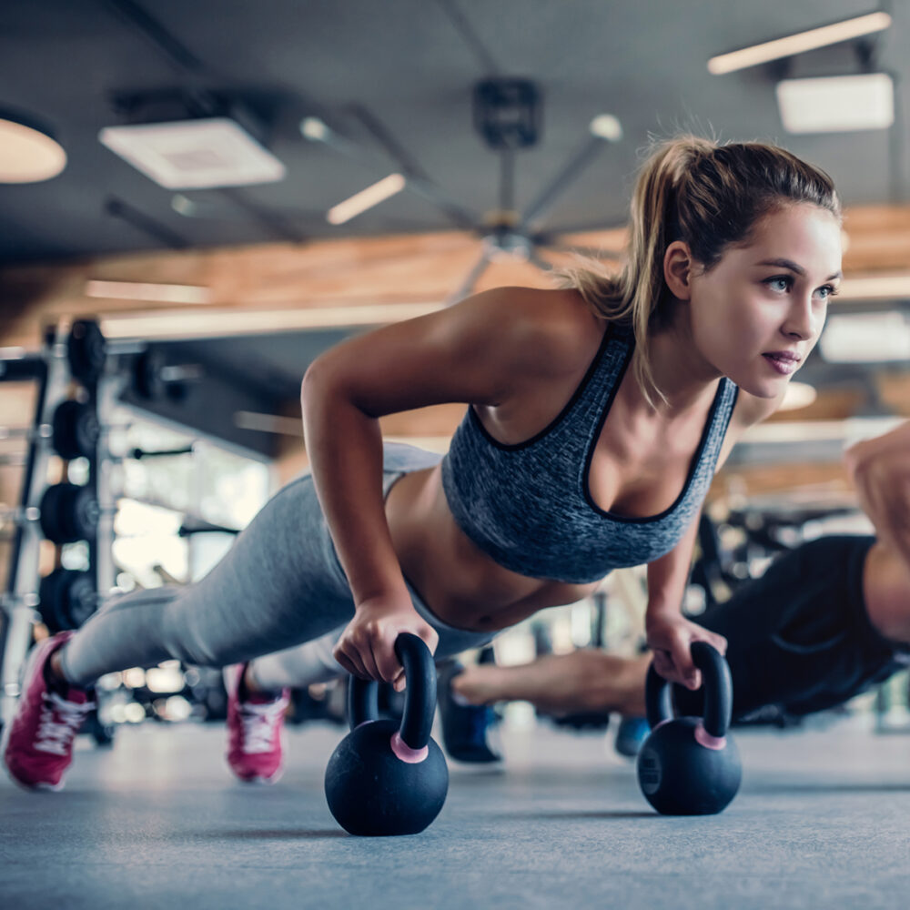 Young couple is working out at gym. Attractive woman and handsome muscular man are training in light modern gym. Doing plank on kettlebell. Push-up on weights.